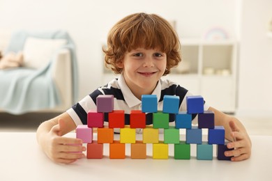 Photo of Little boy stacking colorful cubes at white table indoors