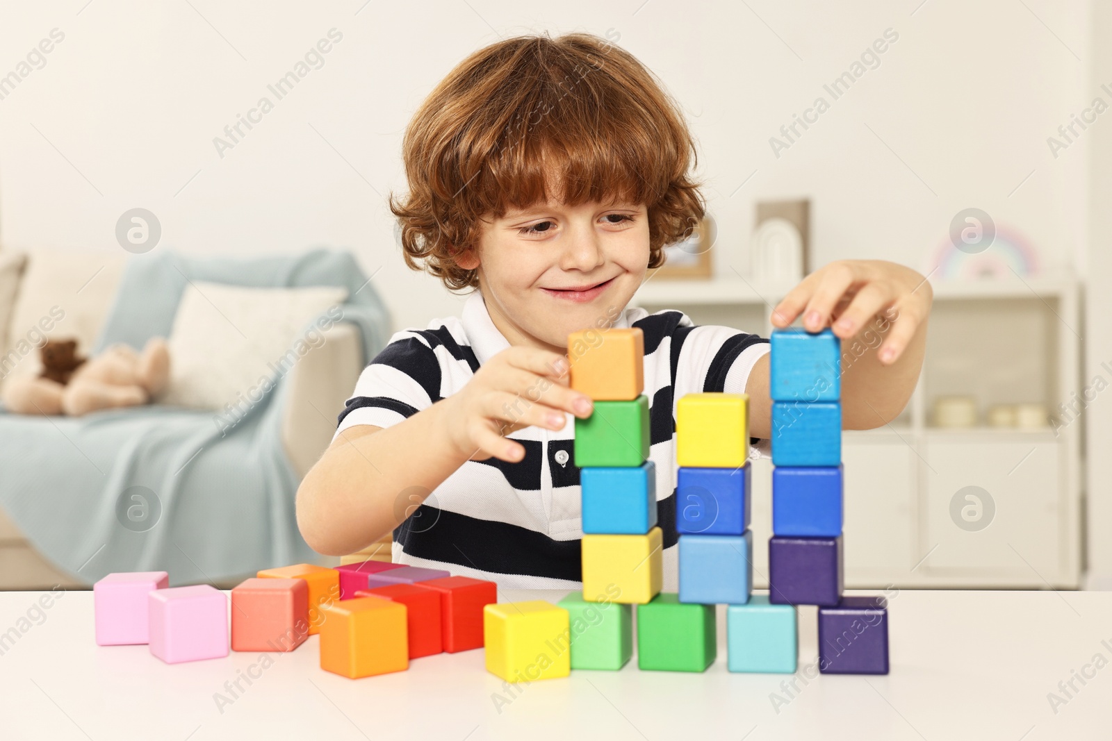 Photo of Little boy stacking colorful cubes at white table indoors