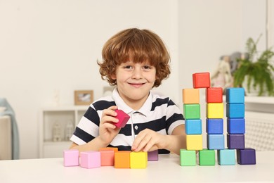 Photo of Little boy stacking colorful cubes at white table indoors