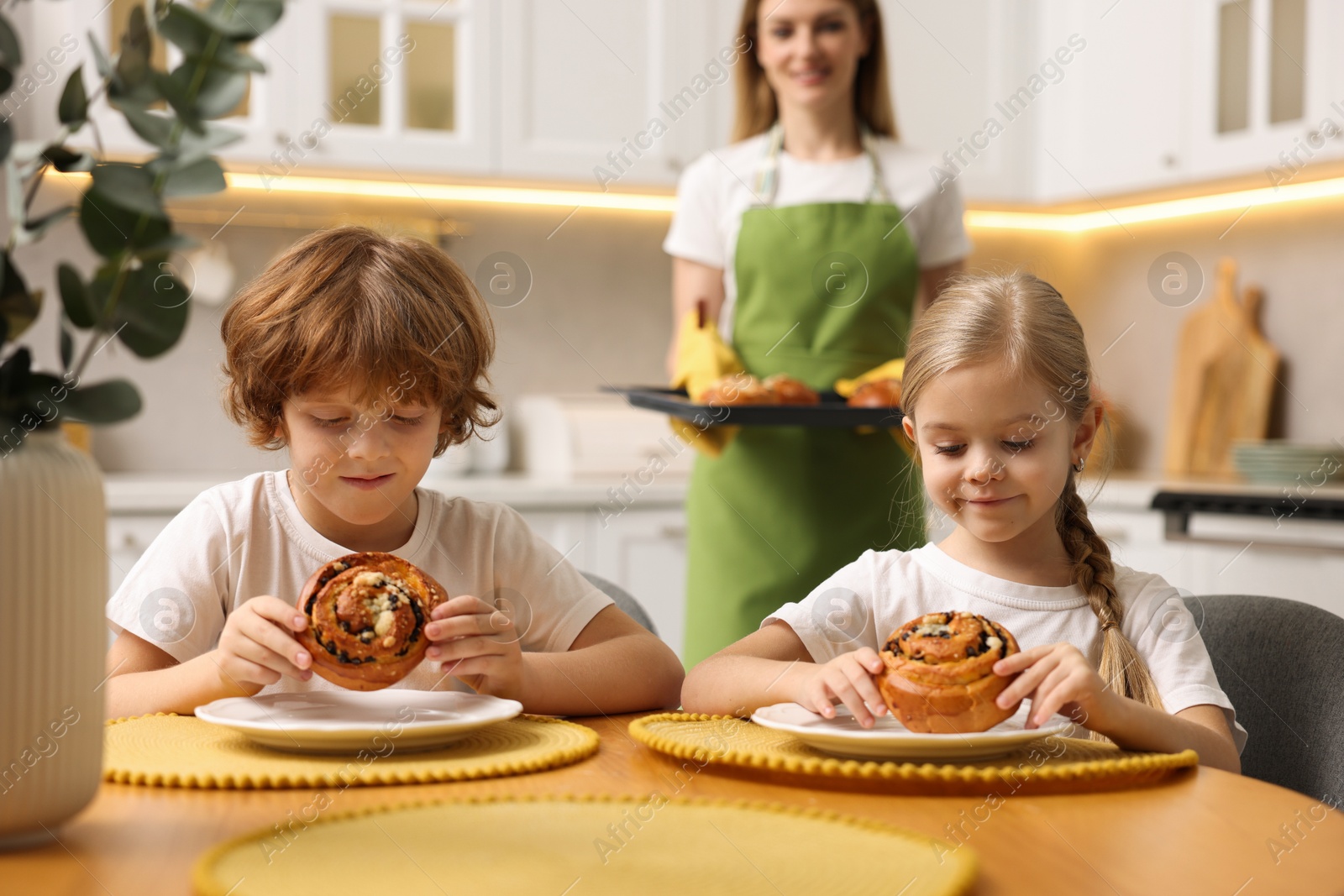 Photo of Cute kids eating tasty buns at table indoors, selective focus. Woman holding baking pan with pastry in kitchen