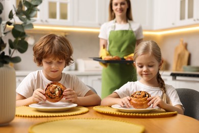 Photo of Cute kids eating tasty buns at table indoors, selective focus. Woman holding baking pan with pastry in kitchen