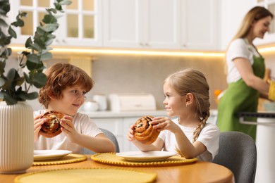 Photo of Cute kids eating tasty buns at table while their mother baking in kitchen, selective focus