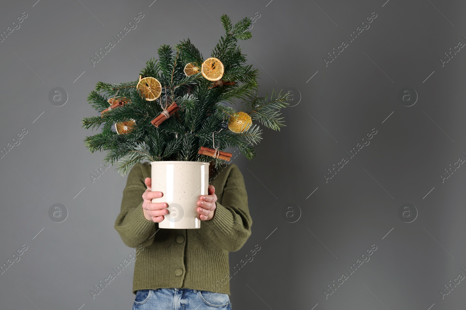 Photo of Woman holding beautiful Christmas composition of fir tree branches decorated with dried orange slices and cinnamon sticks on grey background. Space for text