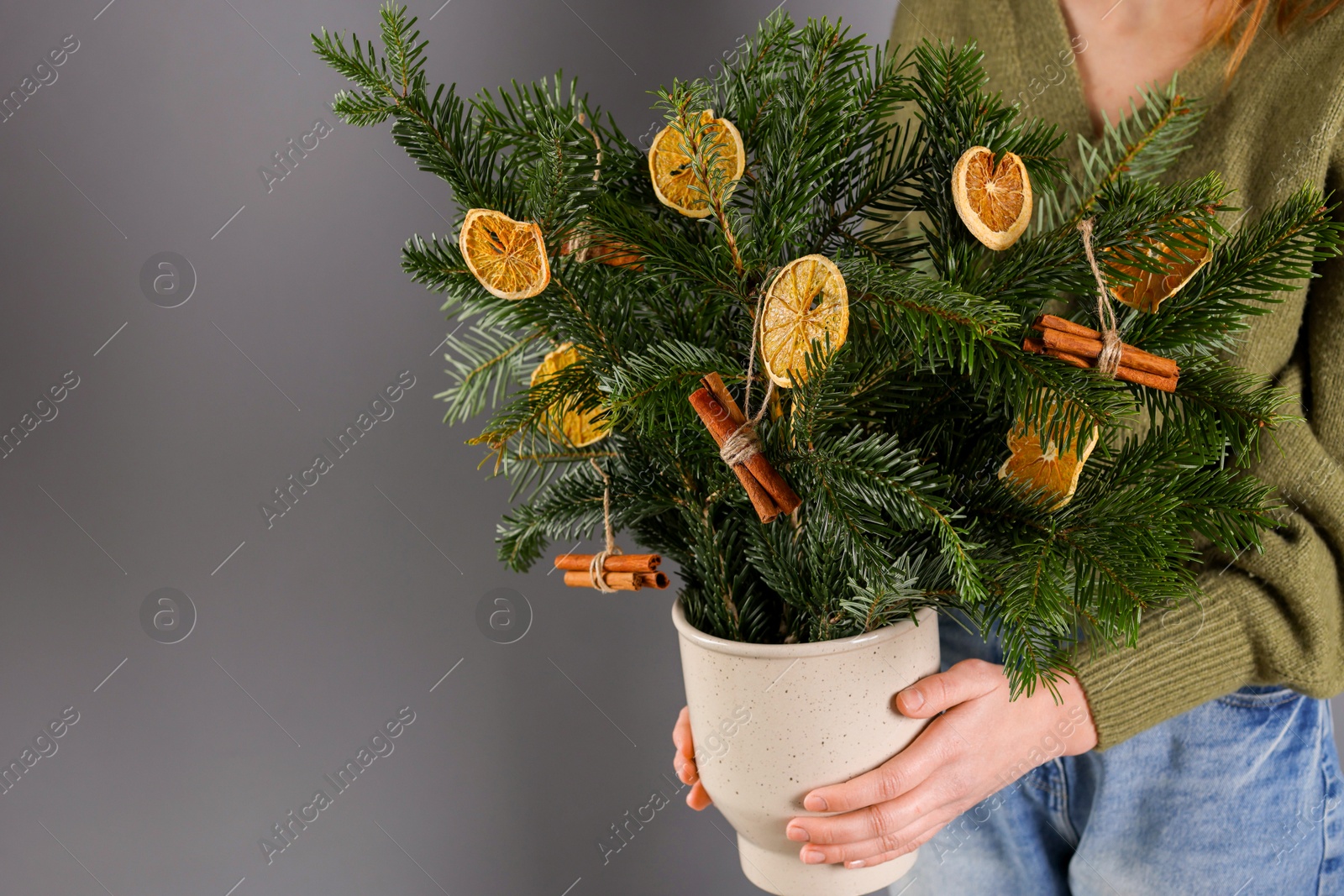 Photo of Woman holding beautiful Christmas composition of fir tree branches decorated with dried orange slices and cinnamon sticks on grey background, closeup. Space for text