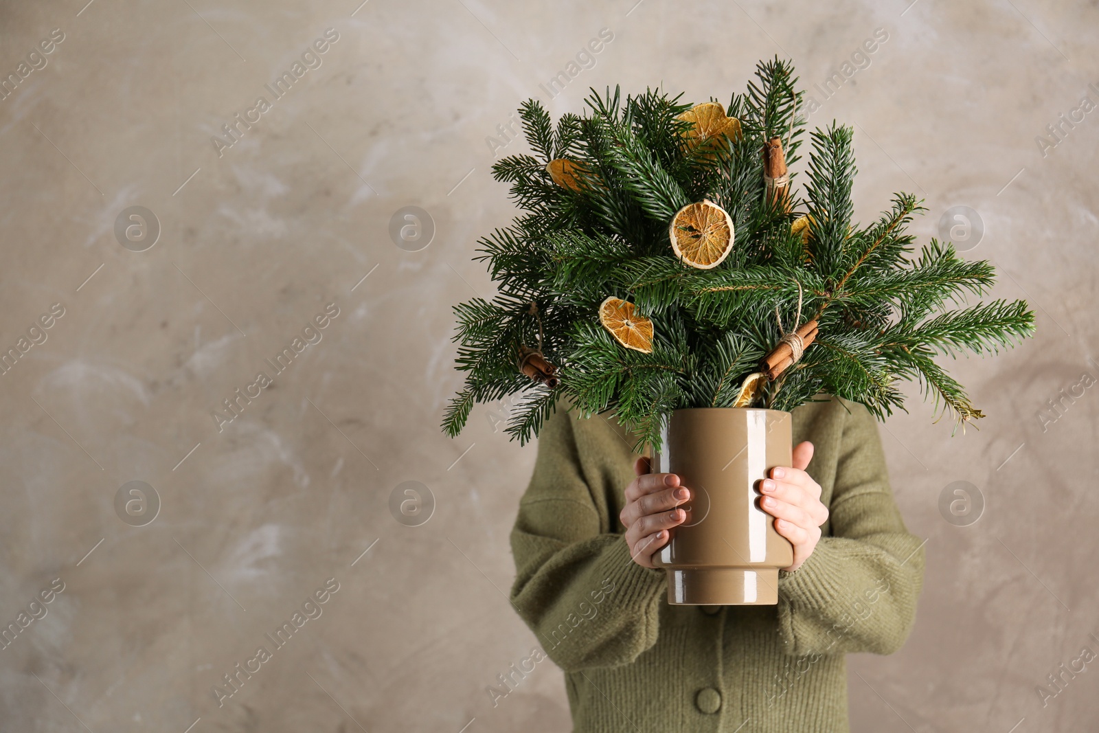 Photo of Woman holding beautiful Christmas composition of fir tree branches decorated with dried orange slices and cinnamon sticks near gray wall, space for text