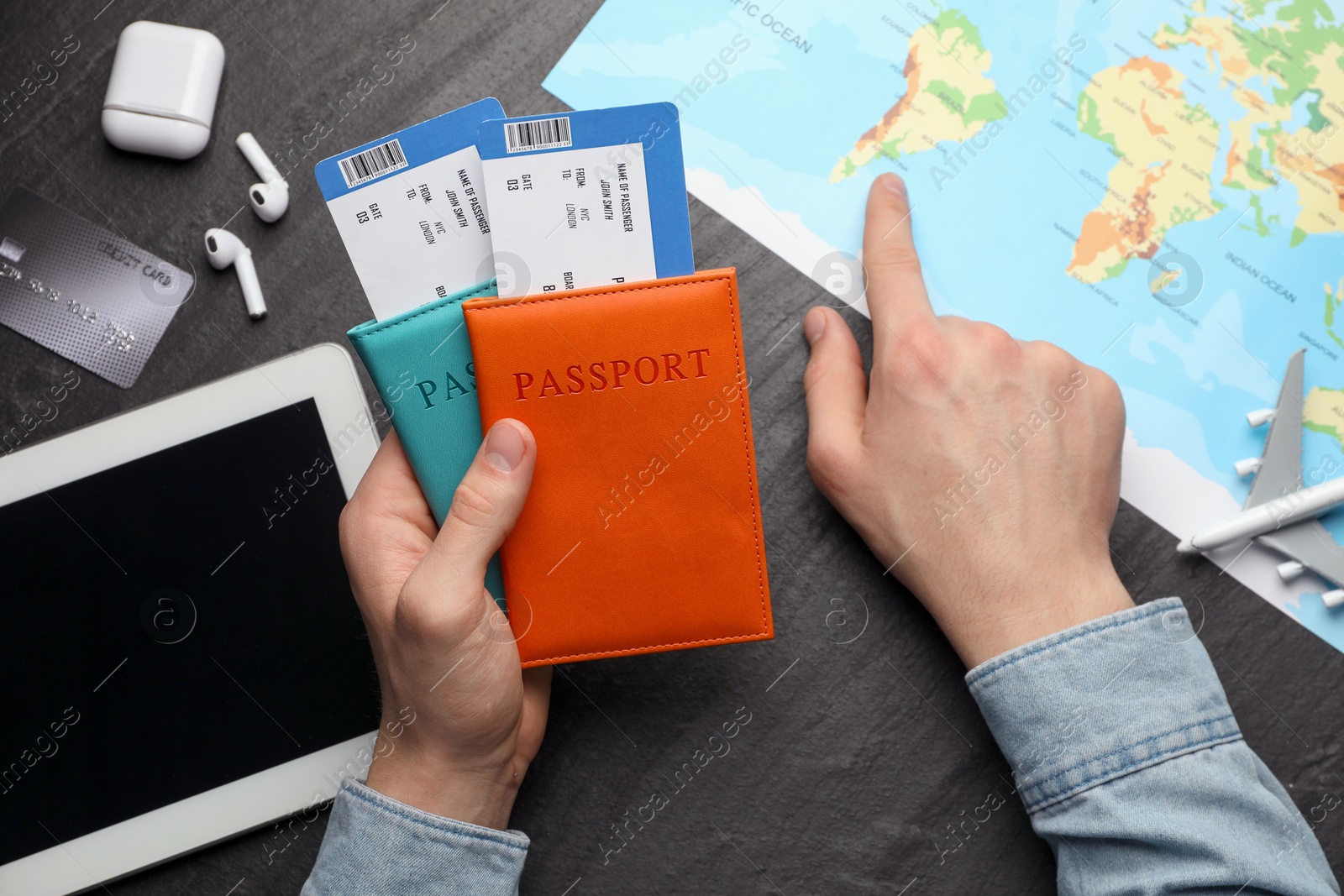 Photo of Travel agency. Man with passports and flight tickets pointing at destination on map at dark textured table, top view