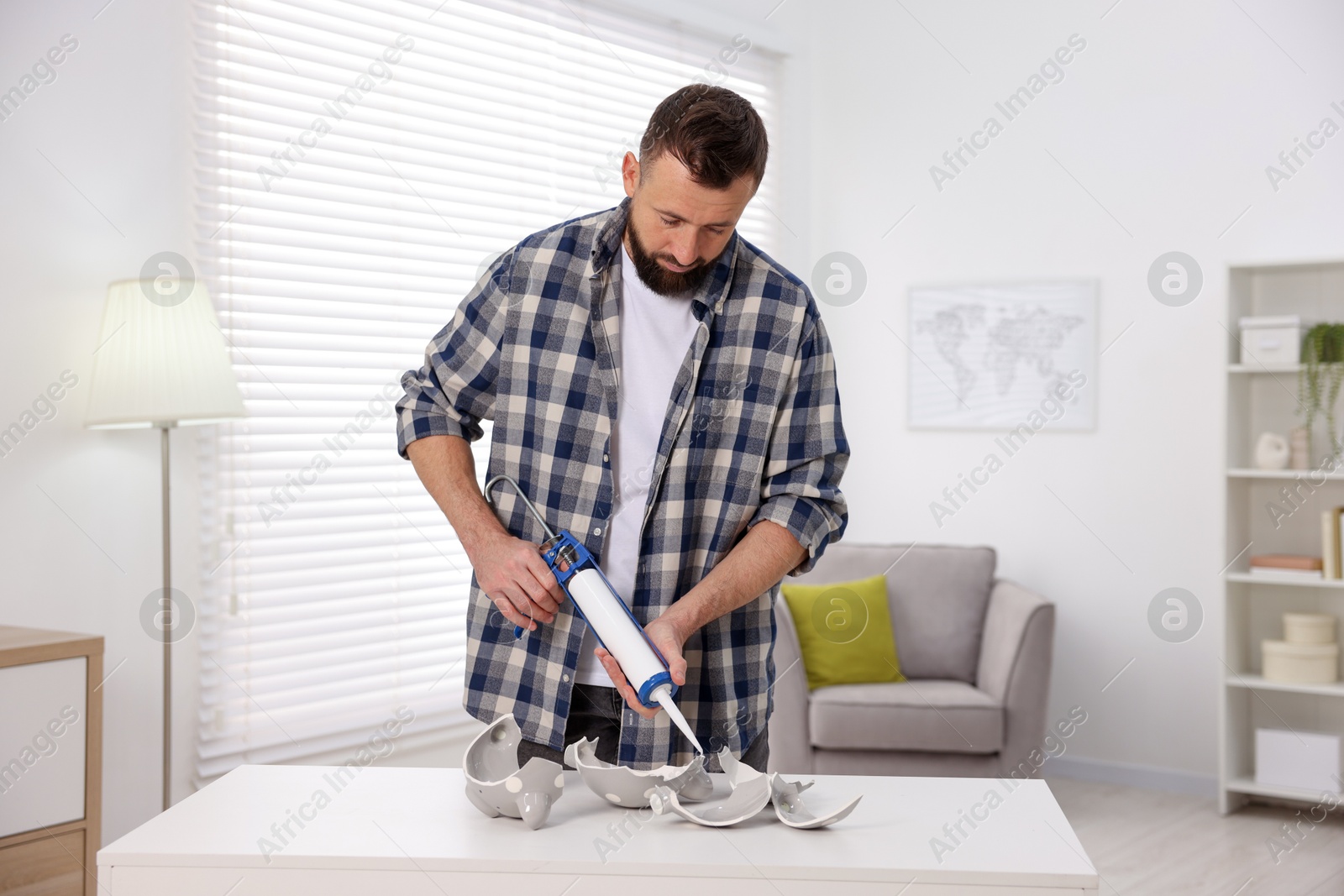 Photo of Man with caulking gun glueing pieces of ceramic piggy bank indoors