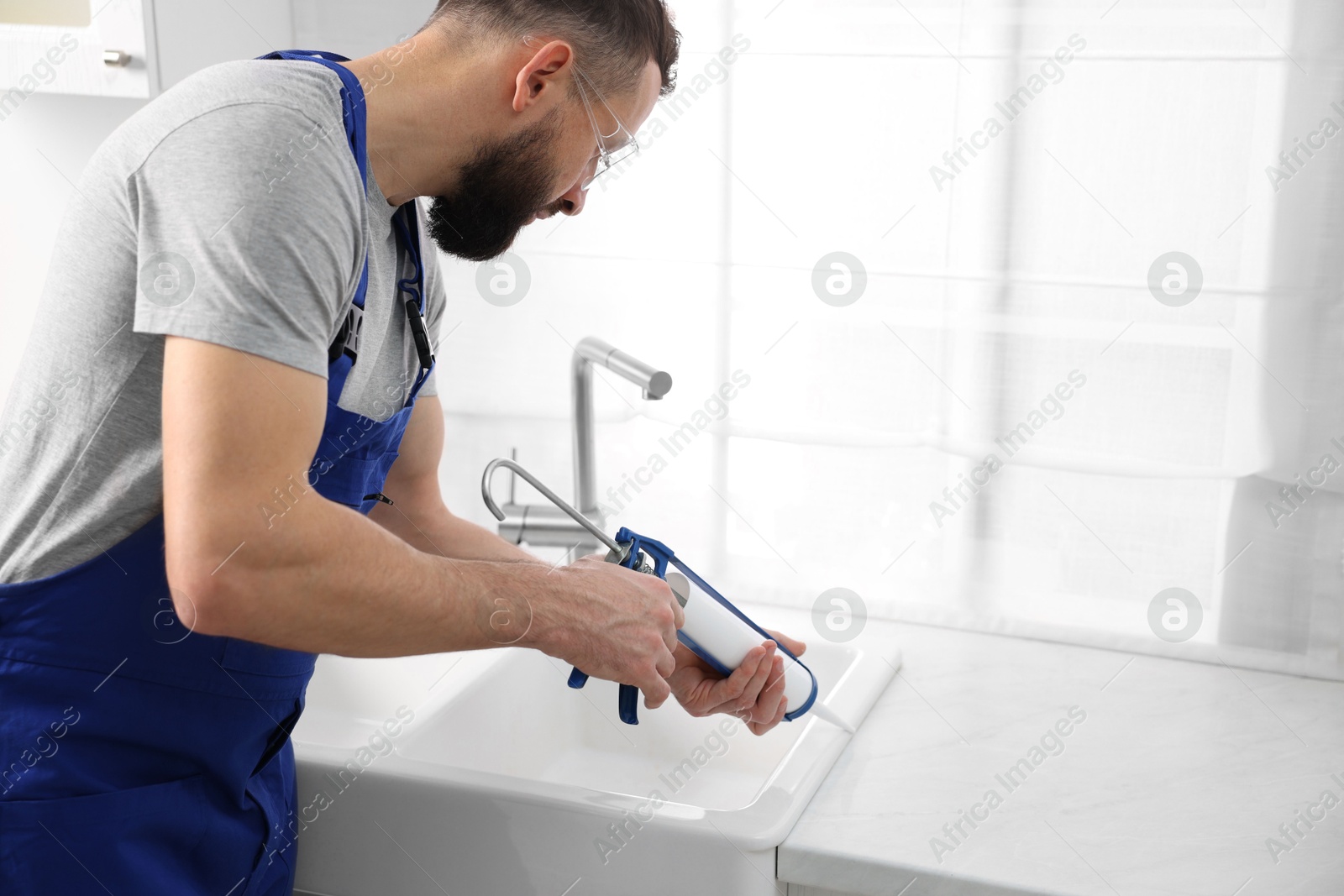 Photo of Worker with caulking gun sealing kitchen sink indoors