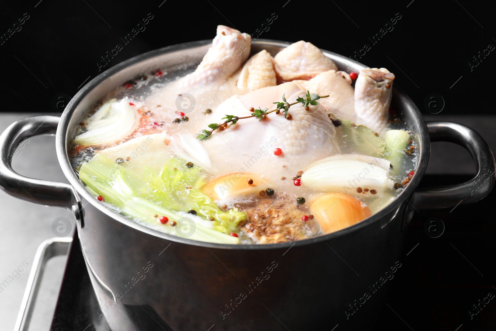 Photo of Whole fresh turkey, cut onion, celery and spices boiling in pot on stove against black background, closeup