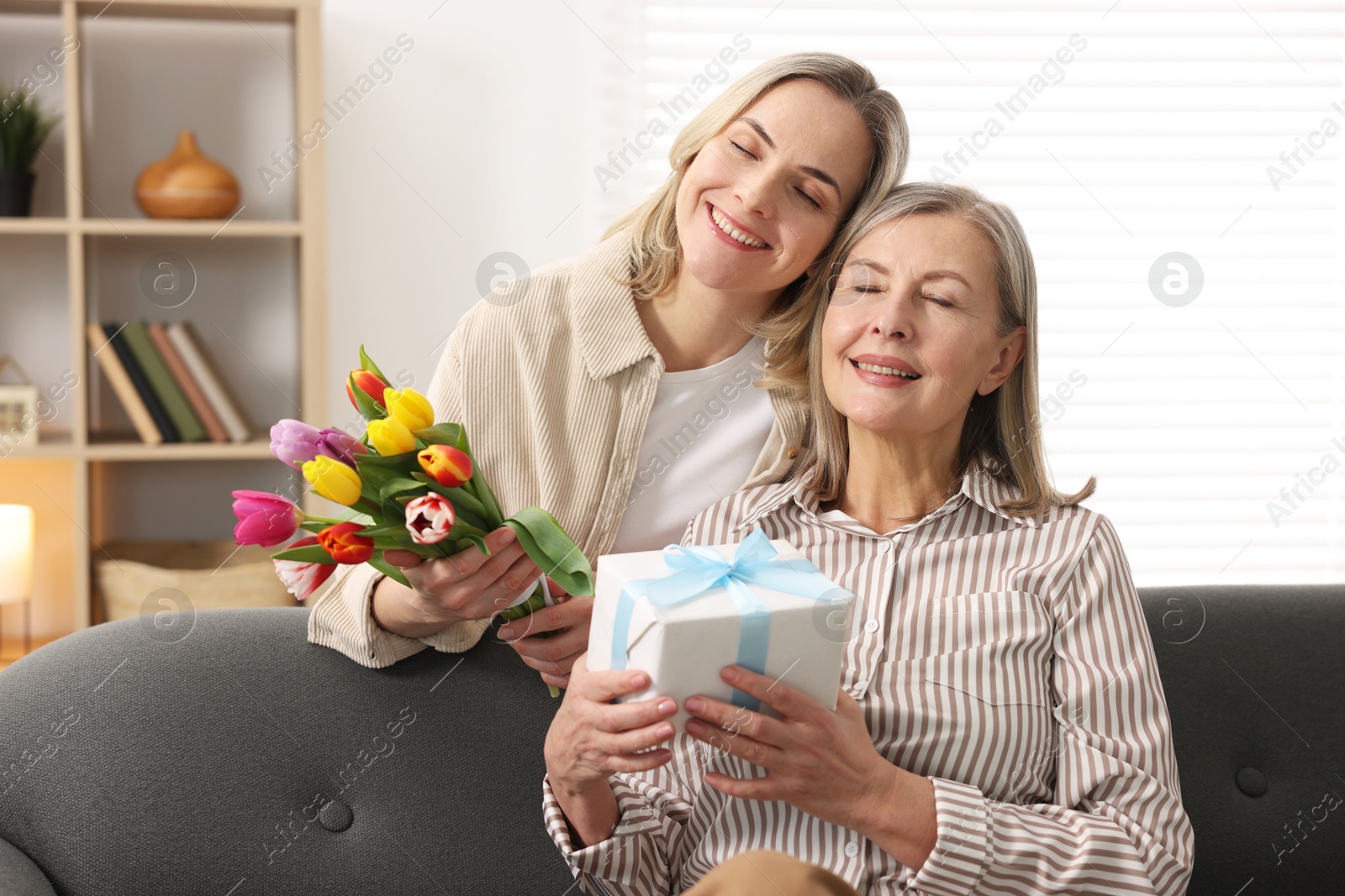 Photo of Smiling daughter congratulating her mom with bouquet of tulips and gift at home. Happy Mother's Day