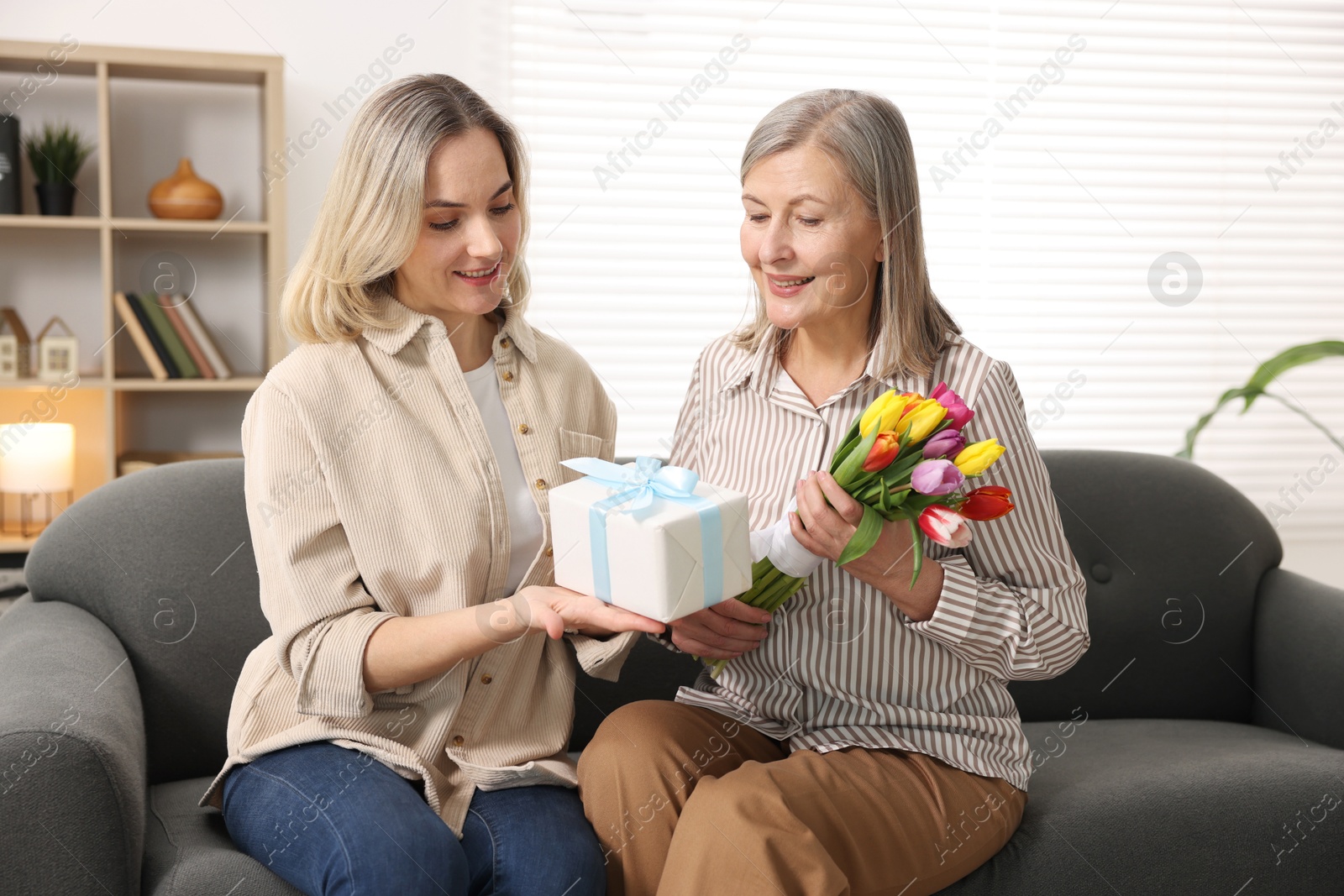 Photo of Smiling daughter congratulating her mom with bouquet of tulips and gift on sofa at home. Happy Mother's Day