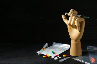 Photo of Drug addiction. Mannequin hand with syringe and pills on black wooden table against dark background, space for text