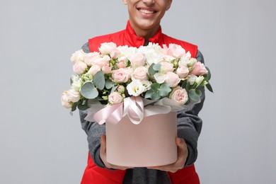 Photo of Smiling delivery man holding gift box with beautiful floral composition on grey background, closeup