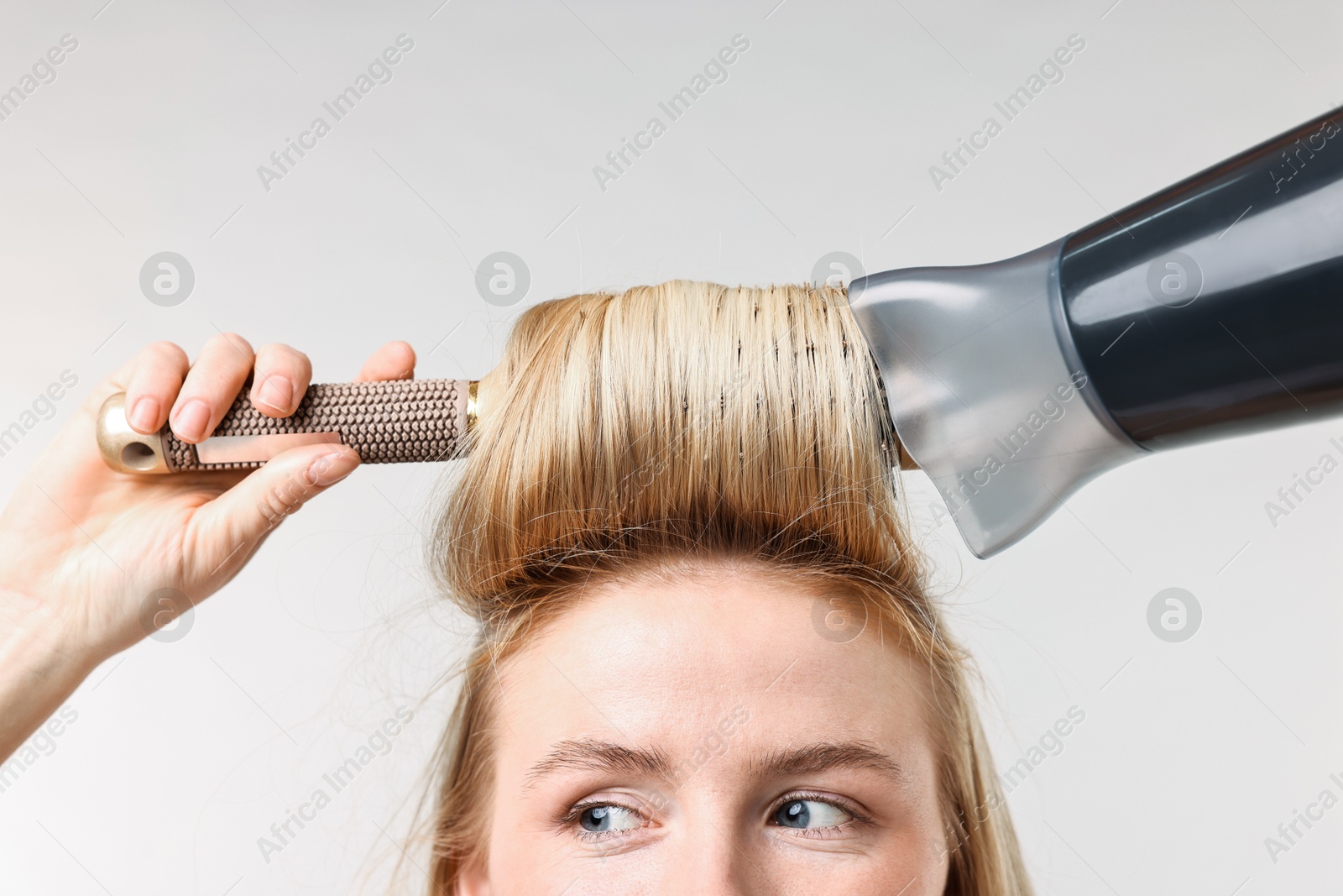 Photo of Beautiful young woman styling her hair with hairdryer and brush on light grey background, closeup