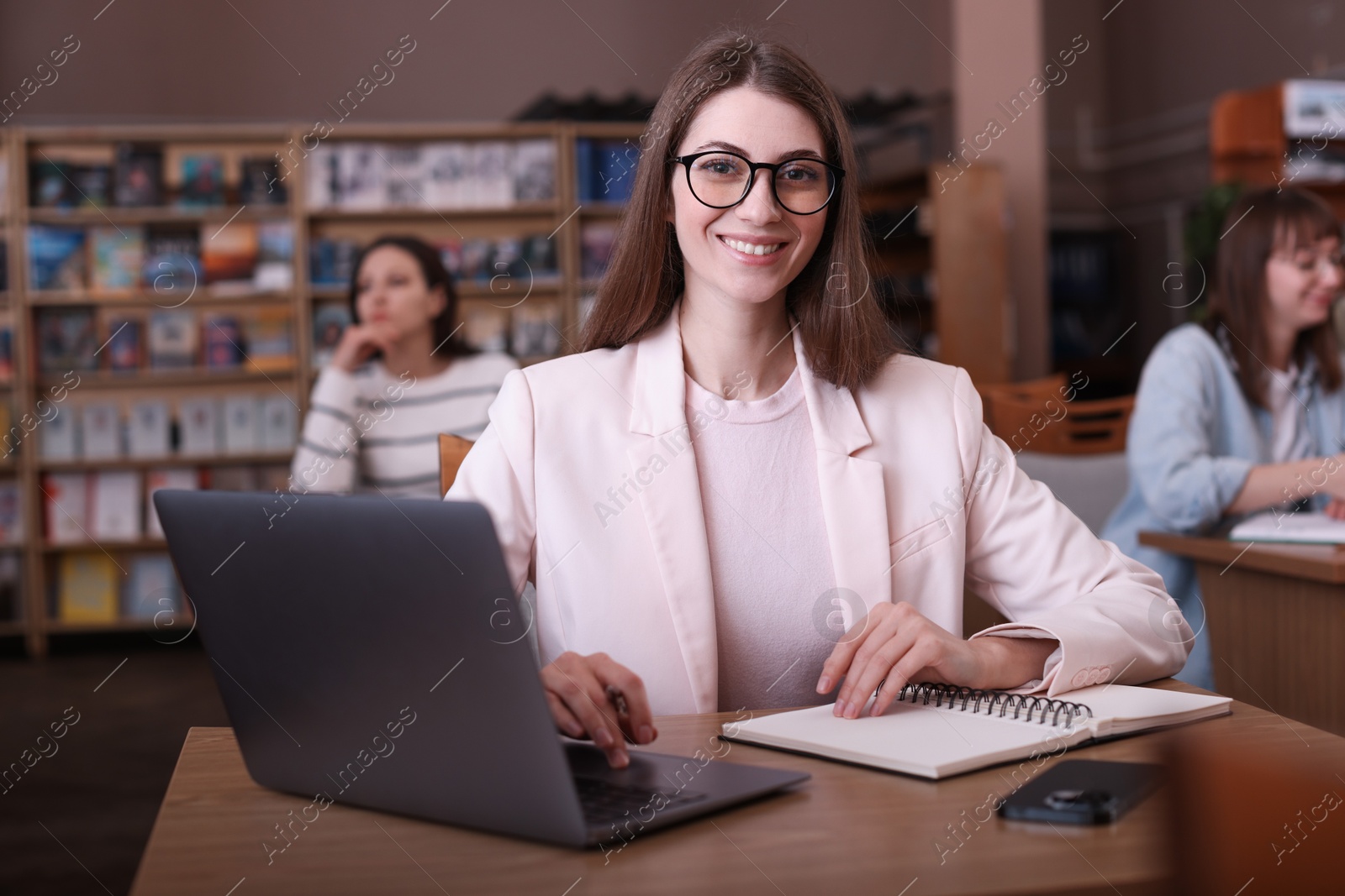 Photo of Portrait of smiling woman with laptop taking notes at desk in public library