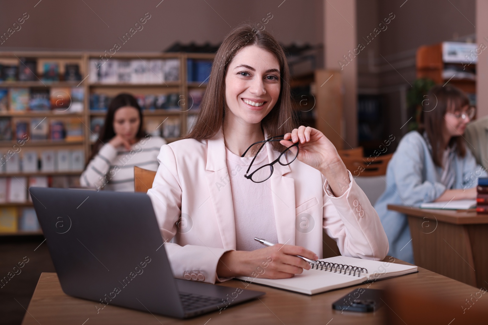 Photo of Portrait of smiling woman with laptop taking notes at desk in public library