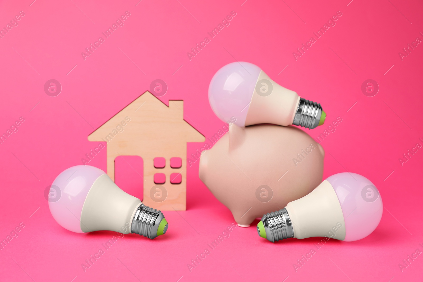 Photo of Piggy bank, light bulbs and wooden house model on pink background. Energy saving concept