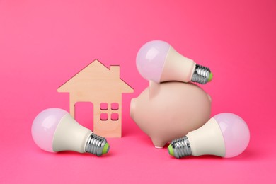 Photo of Piggy bank, light bulbs and wooden house model on pink background. Energy saving concept