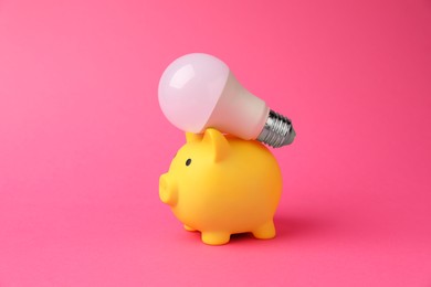 Photo of Piggy bank with light bulb on pink background. Energy saving concept