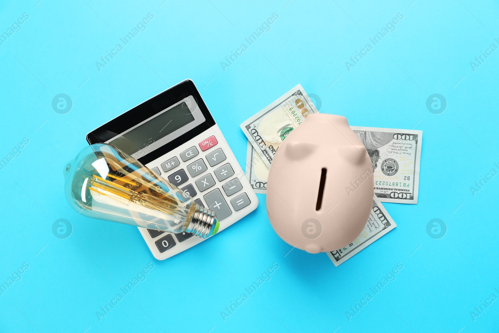 Photo of Piggy bank, dollar banknotes, calculator and lightbulb on light blue background, flat lay. Energy saving concept