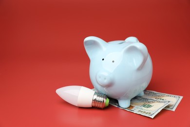 Photo of Piggy bank, dollar banknotes and light bulb on red background, space for text. Energy saving concept