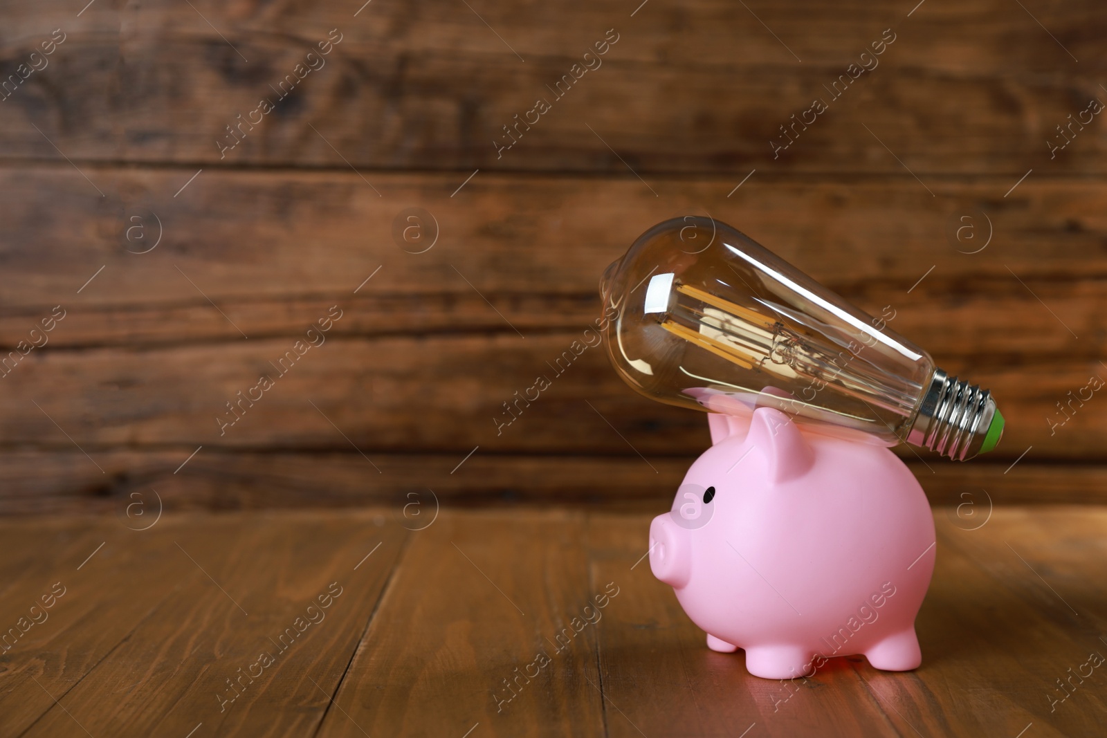 Photo of Piggy bank and light bulb on wooden table, space for text. Energy saving concept