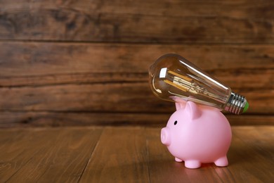 Photo of Piggy bank and light bulb on wooden table, space for text. Energy saving concept