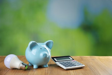 Photo of Piggy bank, calculator and light bulb on wooden table outdoors, space for text. Energy saving concept