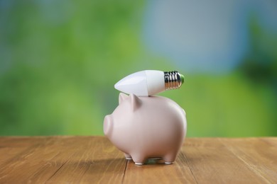 Photo of Piggy bank and light bulb on wooden table outdoors. Energy saving concept