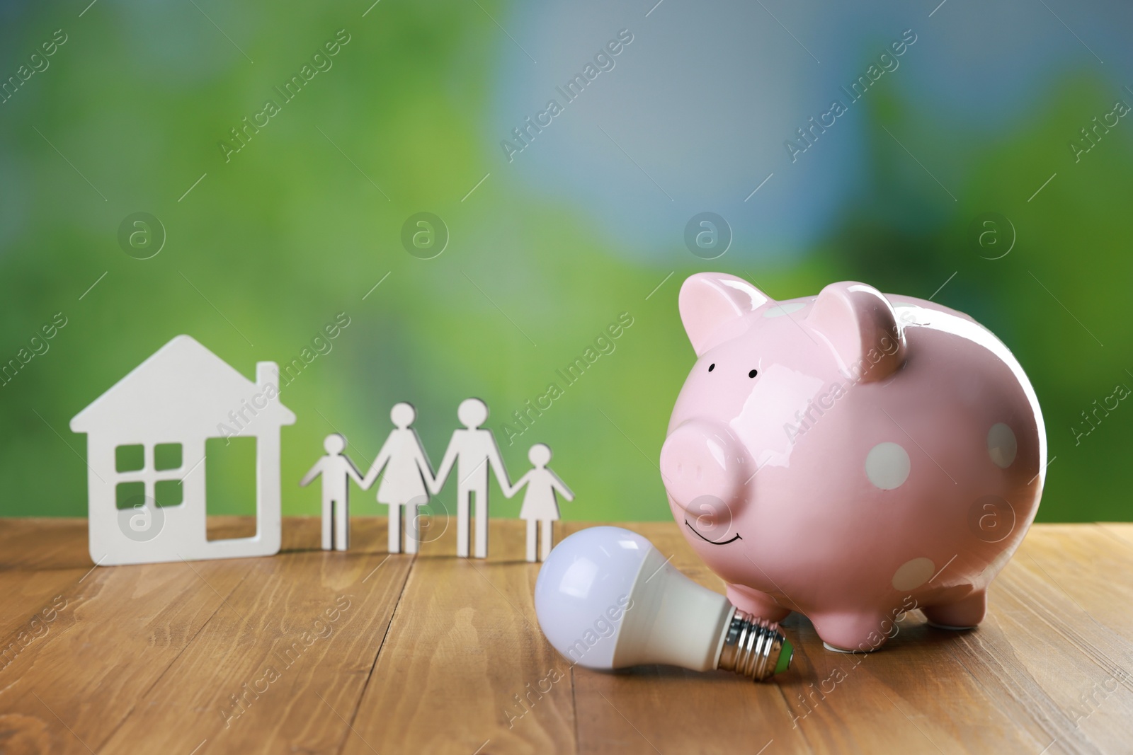 Photo of Piggy bank, house model, figure of family and light bulb on wooden table outdoors. Energy saving concept