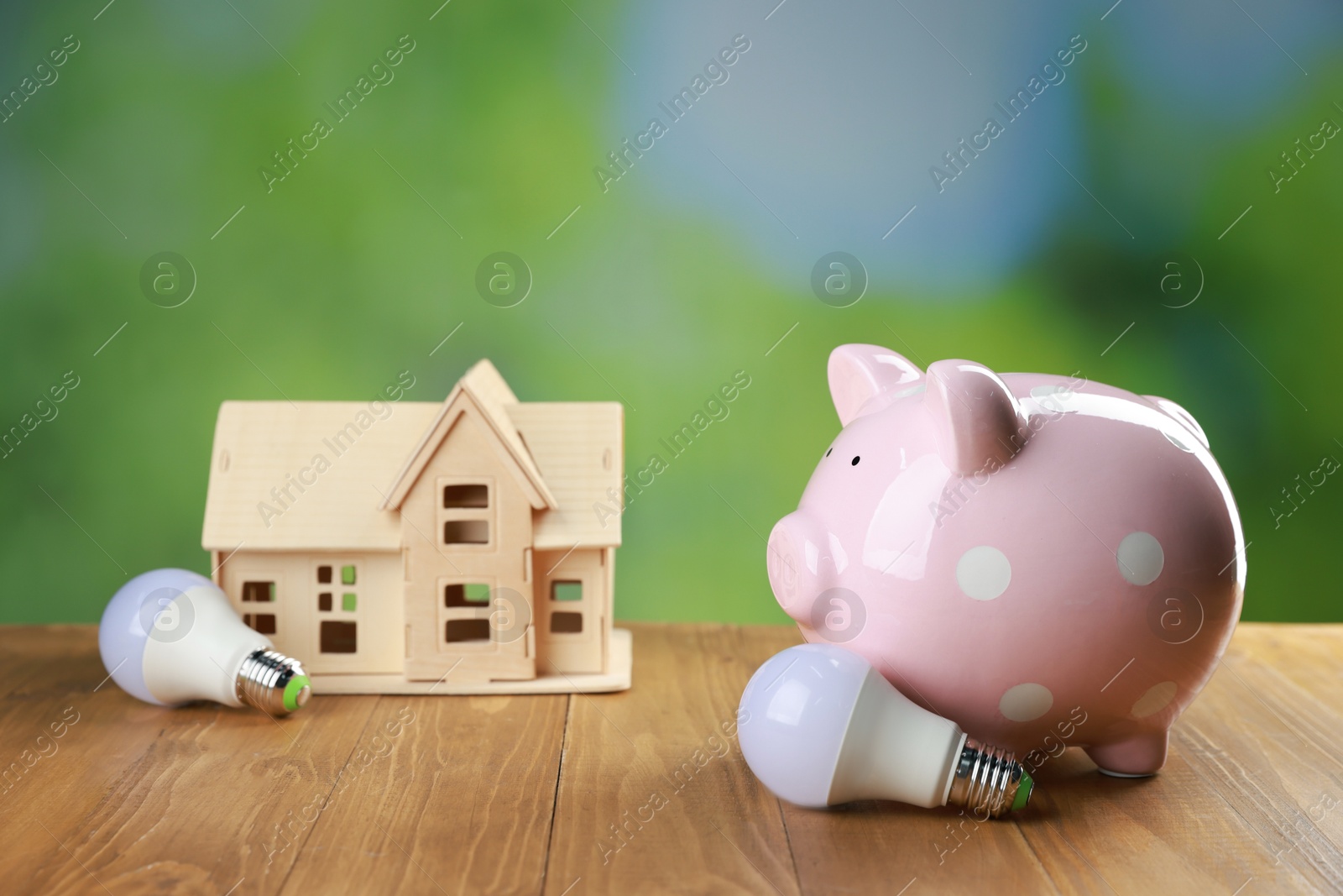 Photo of Piggy bank, house figure and light bulbs on wooden table outdoors. Energy saving concept