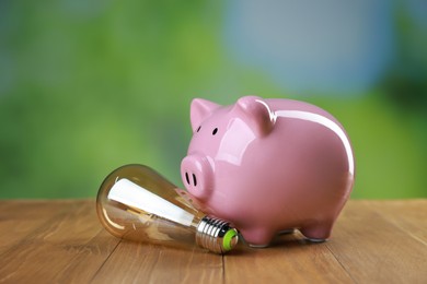 Photo of Piggy bank and light bulb on wooden table outdoors. Energy saving concept