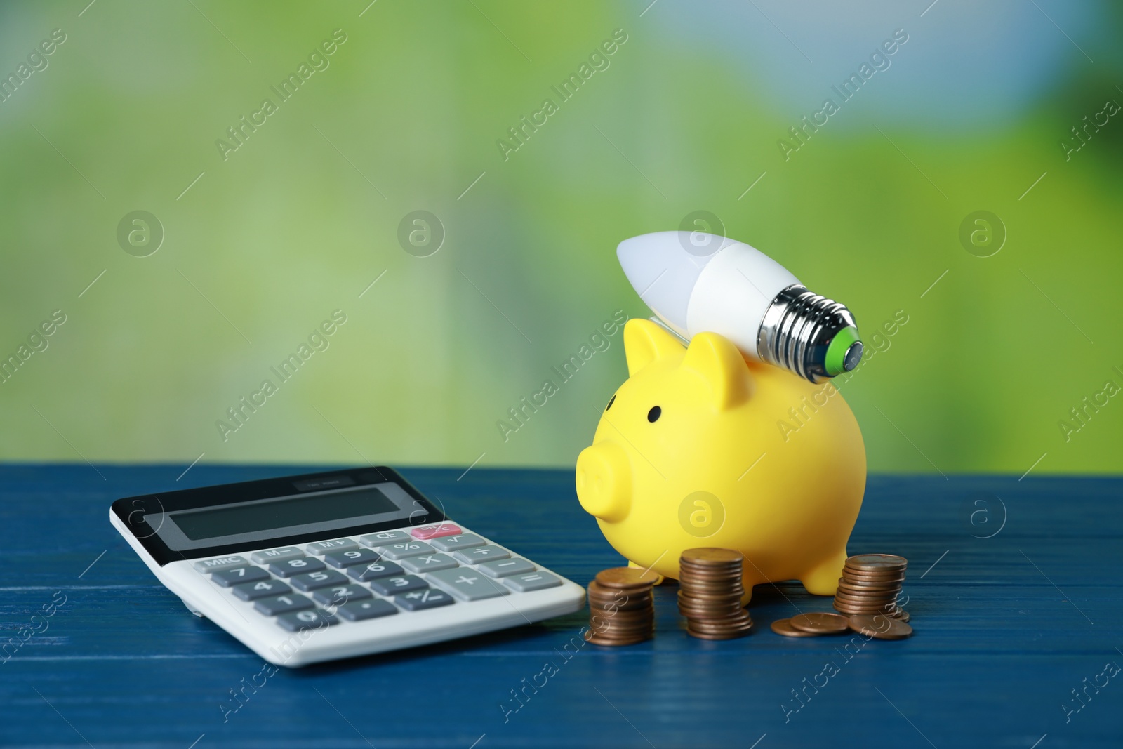 Photo of Piggy bank, calculator, coins and light bulb on blue wooden table outdoors. Energy saving concept