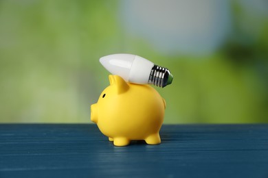 Photo of Piggy bank and light bulb on blue wooden table outdoors. Energy saving concept