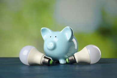 Photo of Piggy bank and light bulbs on blue wooden table outdoors. Energy saving concept