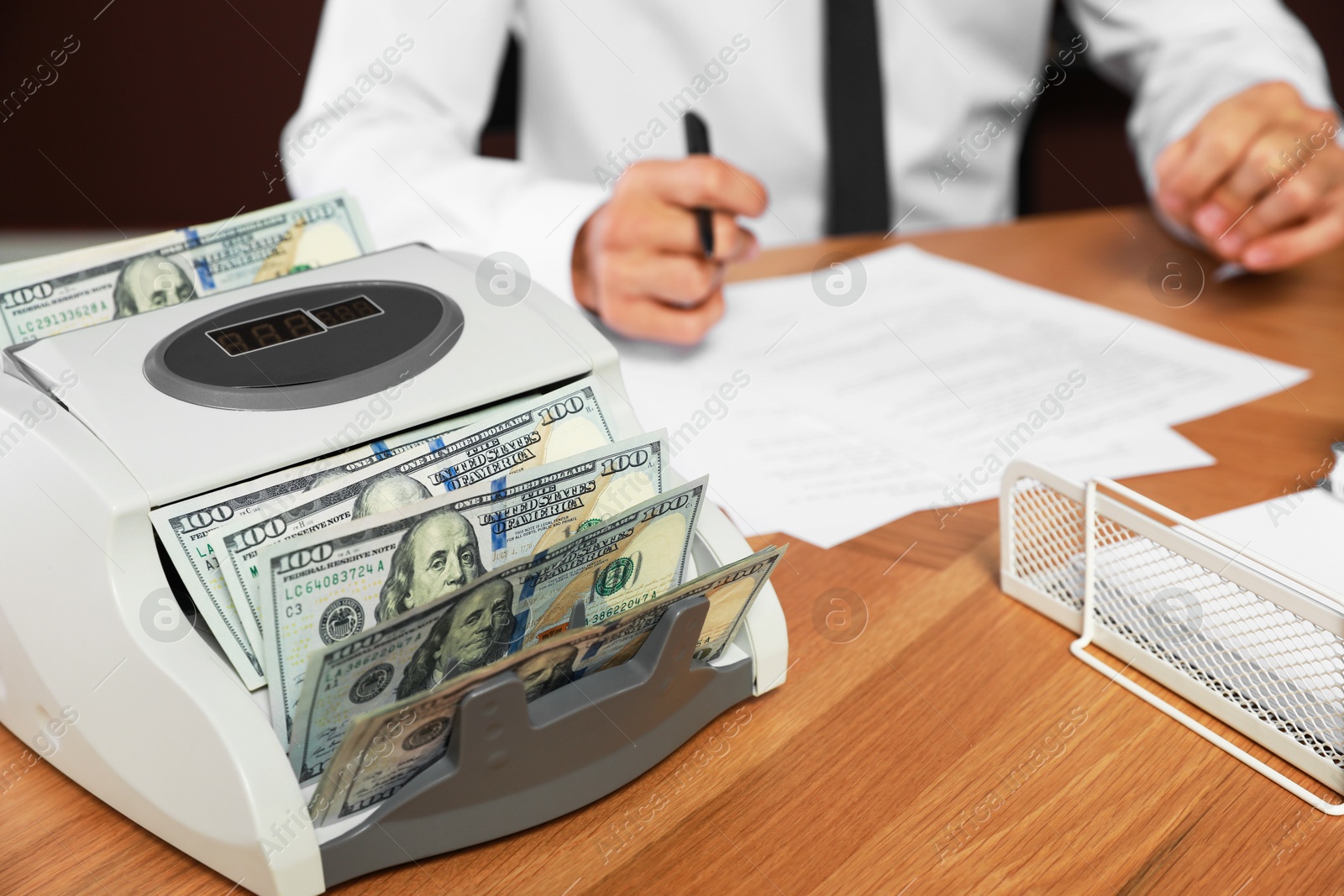 Photo of Modern banknote counter with money and blurred view of man working at wooden table indoors