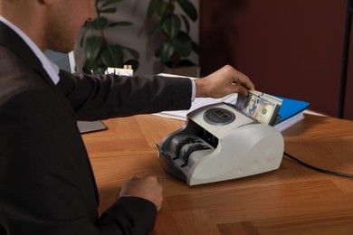 Photo of Man putting money into banknote counter at wooden table indoors, closeup