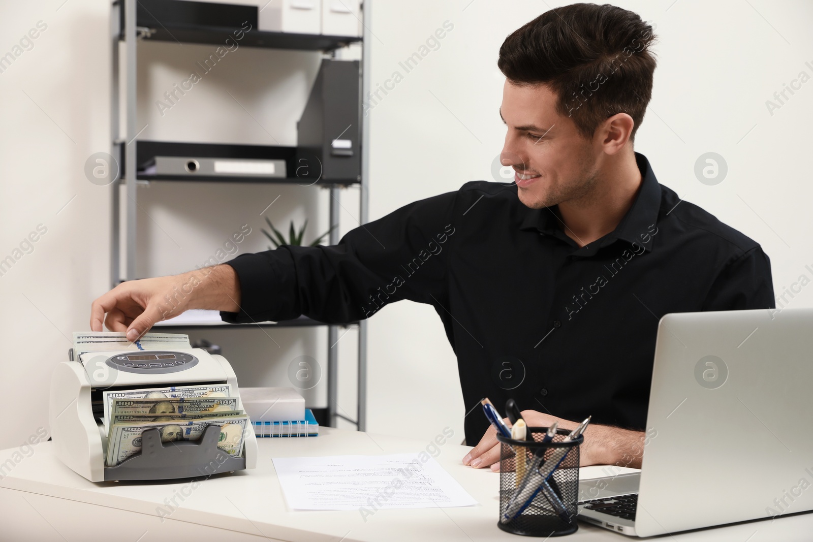 Photo of Man putting money into banknote counter at white table indoors