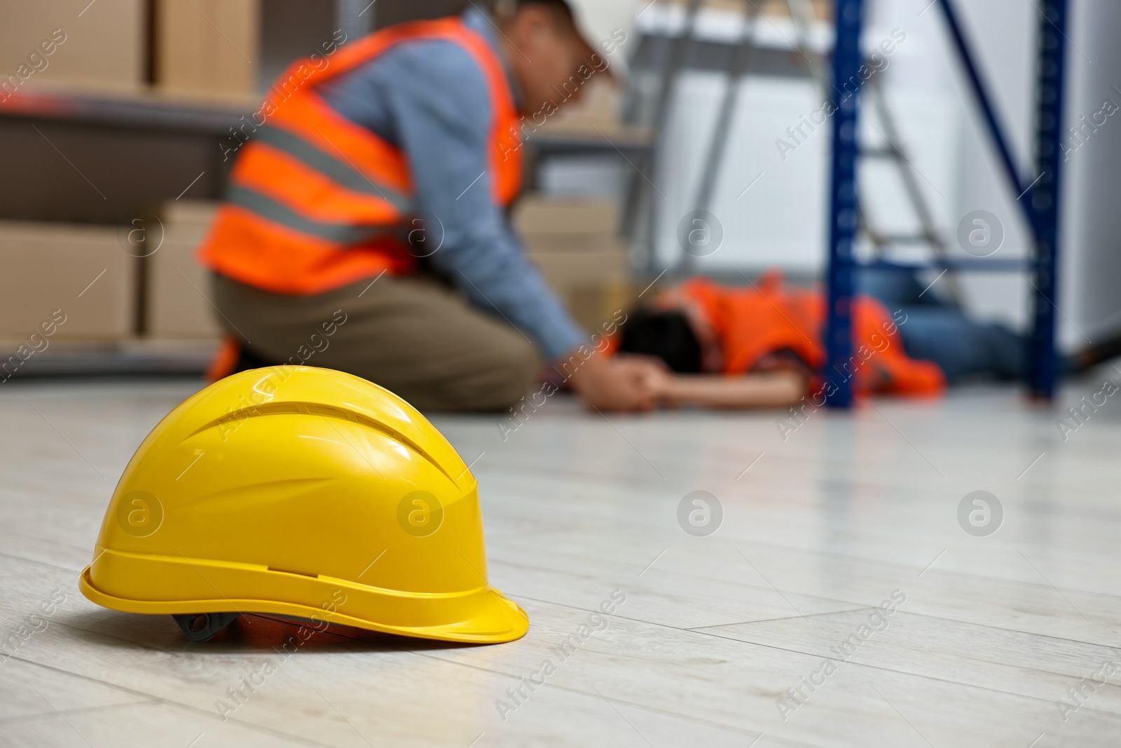 Photo of Accident at work. Hardhat on floor and man checking pulse of his unconscious colleague in warehouse, selective focus