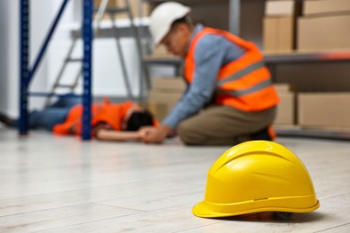 Photo of Accident at work. Hardhat on floor and man checking pulse of his unconscious colleague in warehouse, selective focus