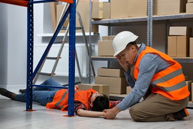 Photo of Accident at work. Man checking pulse of his unconscious colleague in warehouse