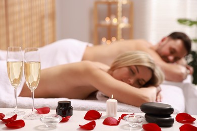 Photo of Couple lying on massage tables in spa salon, focus of petals, burning candle and glasses of champagne