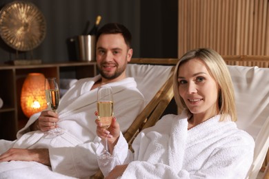 Photo of Happy couple in bathrobes with glasses of champagne relaxing in spa salon, selective focus