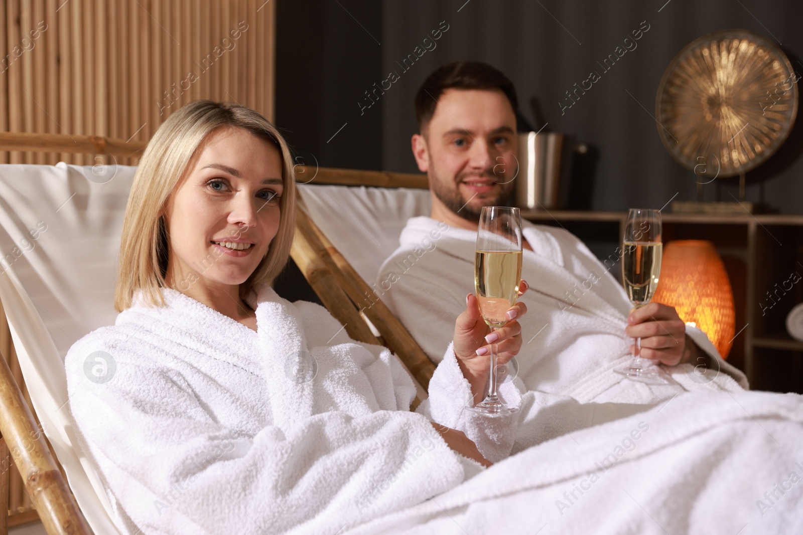 Photo of Happy couple in bathrobes with glasses of champagne relaxing in spa salon, selective focus