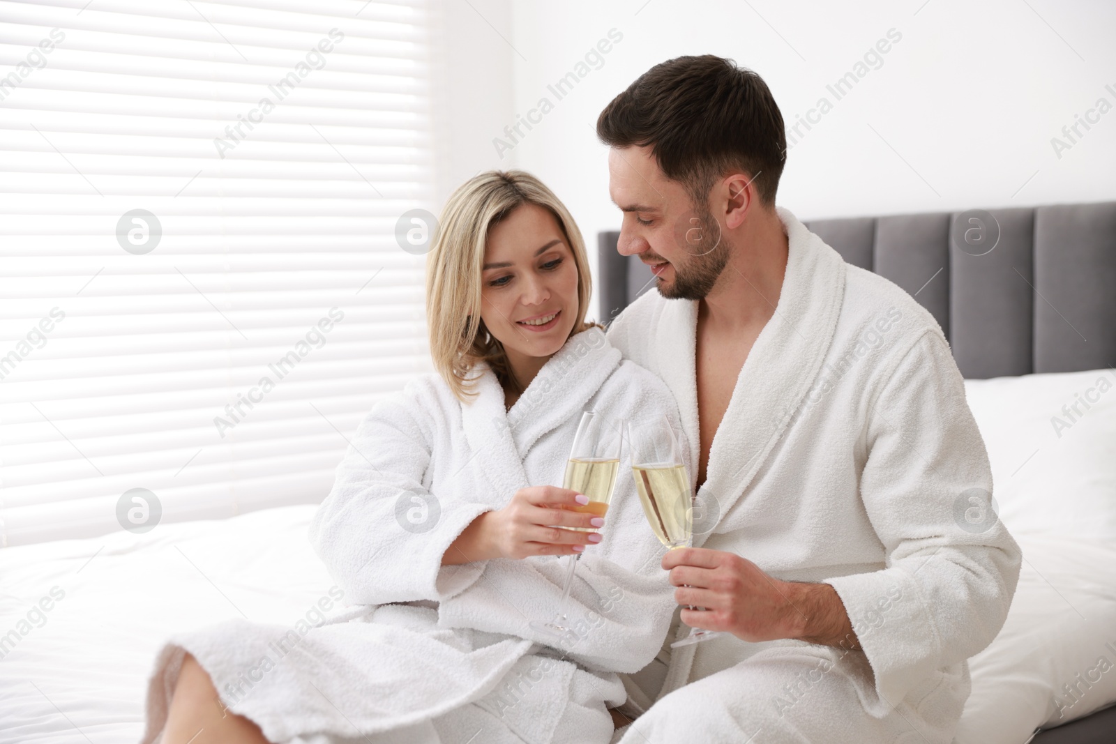 Photo of Spa. Happy couple in bathrobes with glasses of champagne sitting on bed indoors