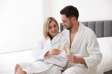 Photo of Spa. Happy couple in bathrobes with glasses of champagne sitting on bed indoors