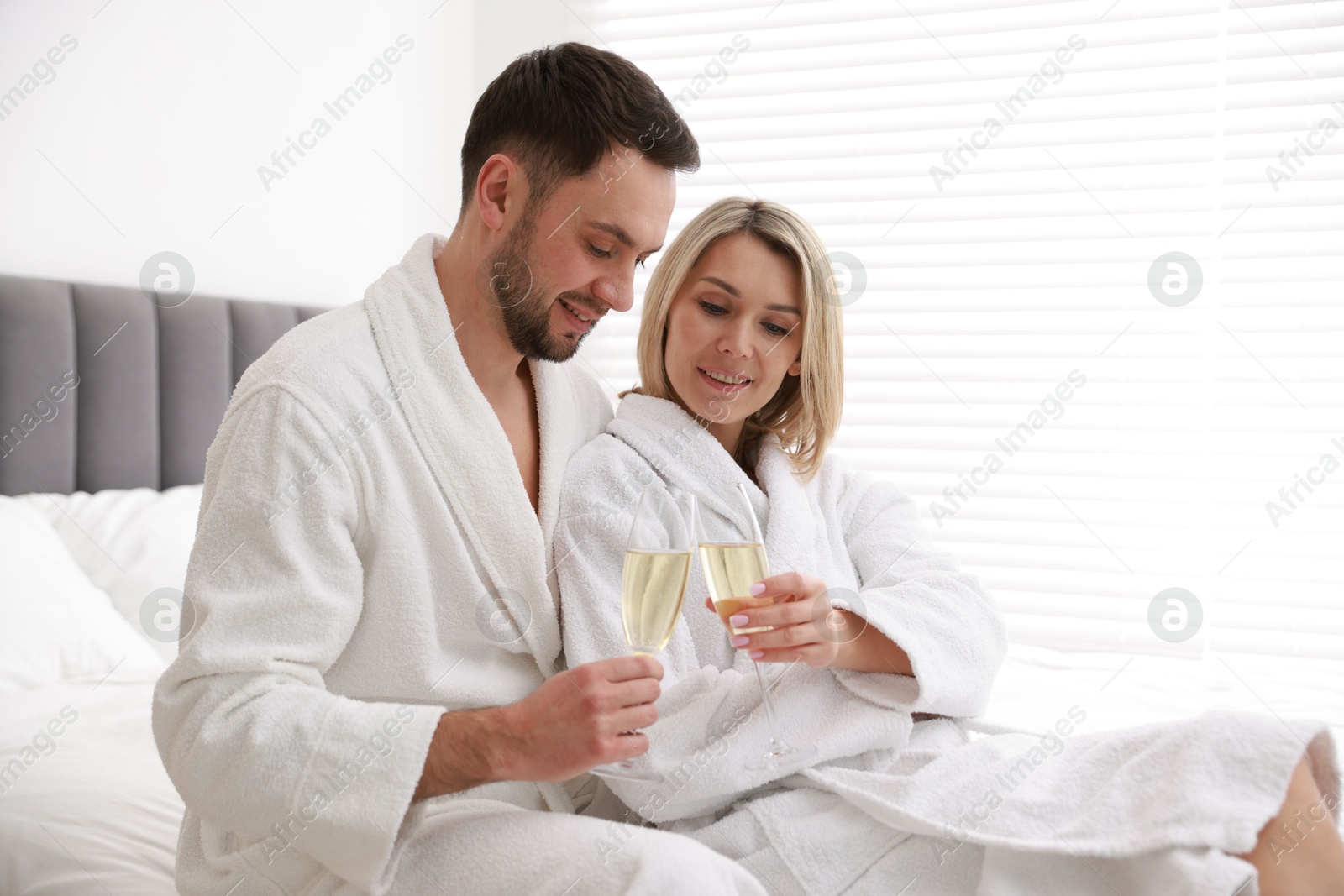 Photo of Spa. Happy couple in bathrobes with glasses of champagne sitting on bed indoors