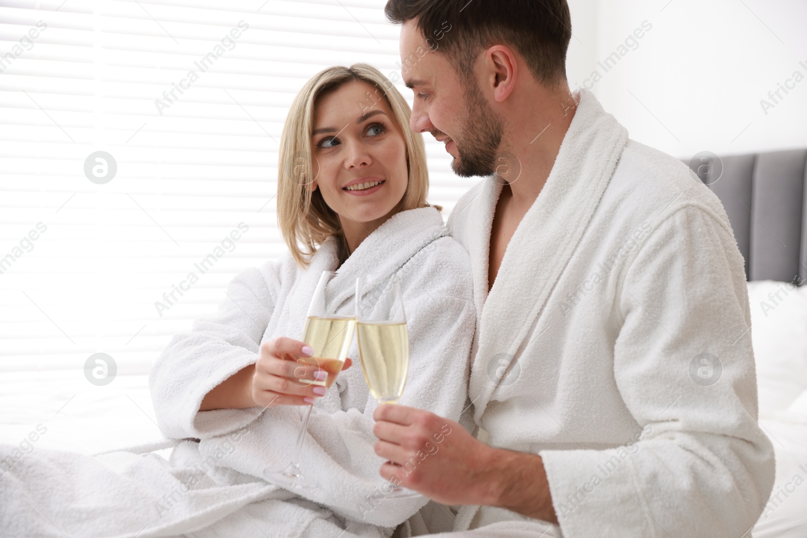 Photo of Spa. Happy couple in bathrobes with glasses of champagne sitting on bed indoors