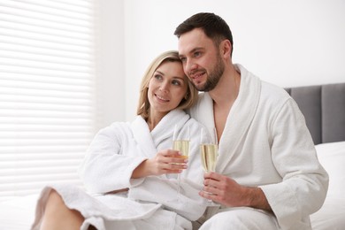 Photo of Spa. Happy couple in bathrobes with glasses of champagne sitting on bed indoors