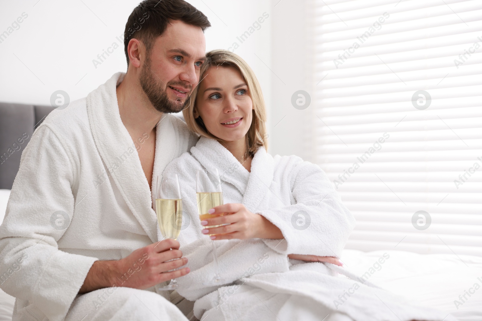 Photo of Spa. Happy couple in bathrobes with glasses of champagne sitting on bed indoors, space for text